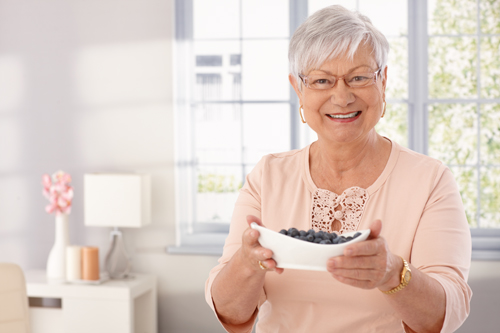 Elderly lady offering a bowl of blueberry, smiling, looking at camera.