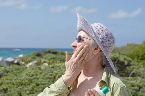A woman wearing a sun hat, long sleeve shirt and sunglasses applies sunscreen on a bright, sunny day.