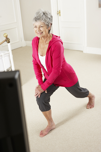 Senior Woman Exercising Whilst Watching Fitness DVD On Television In Bedroom.