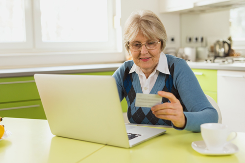 Cheerful senior woman is sitting at the kitchen table and using laptop to do some online shopping and make payment with credit card.