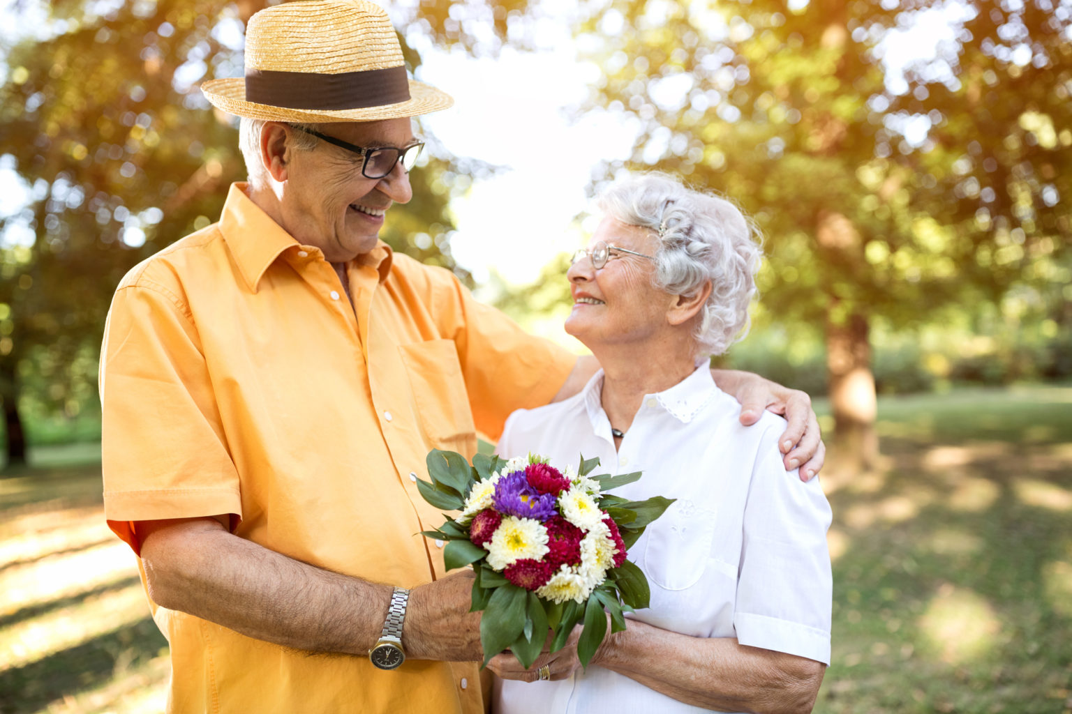 A senior couple looks happily at each other. The woman is holding flowers.