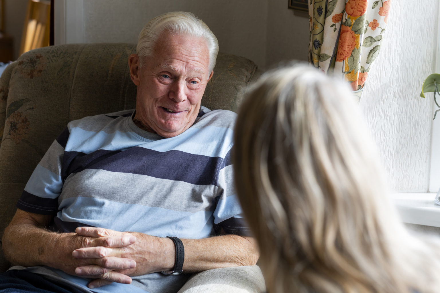 Nurse conversing with a senior patient in the North East of England while he is sitting in a chair in his living room.