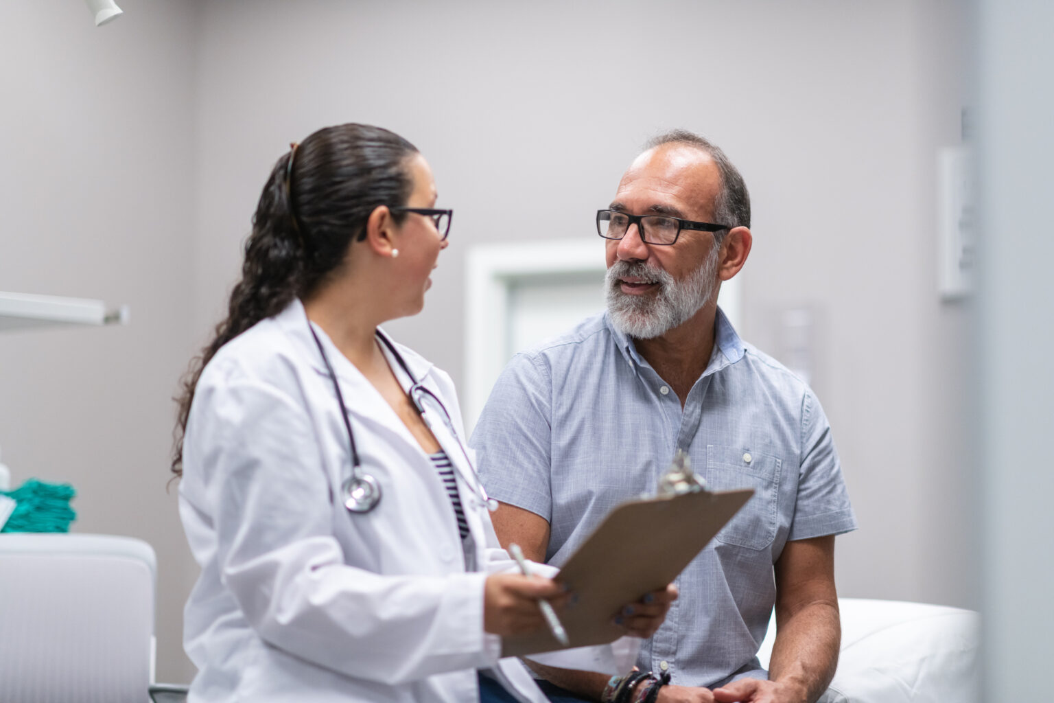 A female doctor of Hispanic descent is consulting a senior male patient. The patient and doctor are sitting facing each other in a medical clinic examination room. The doctor is showing the patient test results on a clipboard.