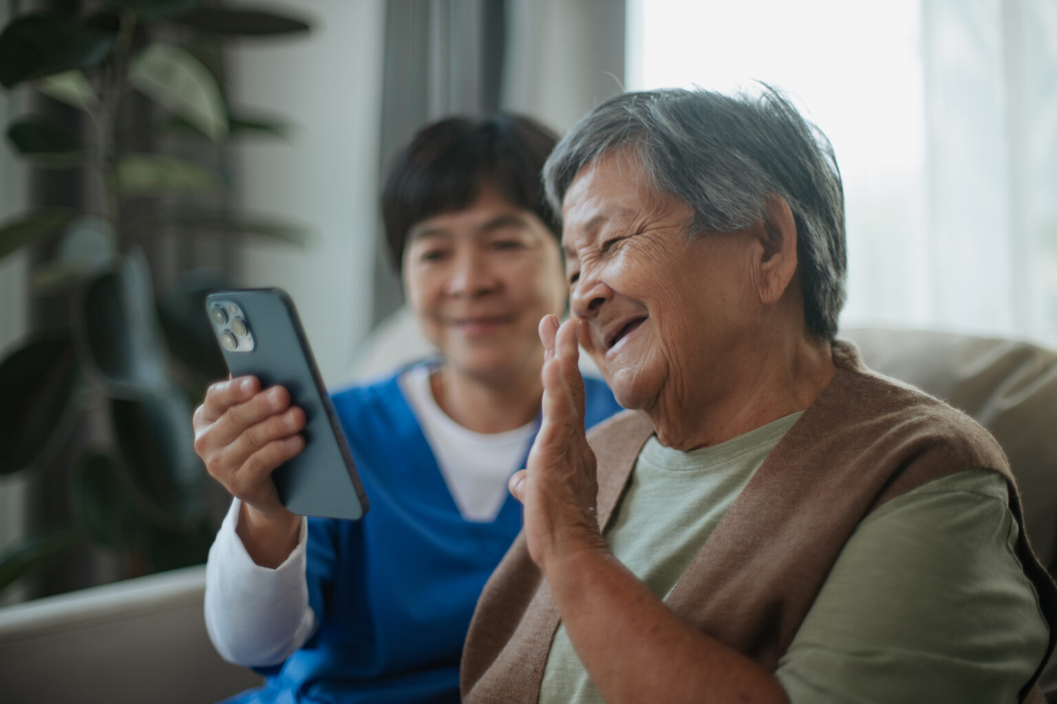 Asian mature caregiver assisting elderly woman making a video call to her family while relaxing on sofa at home.