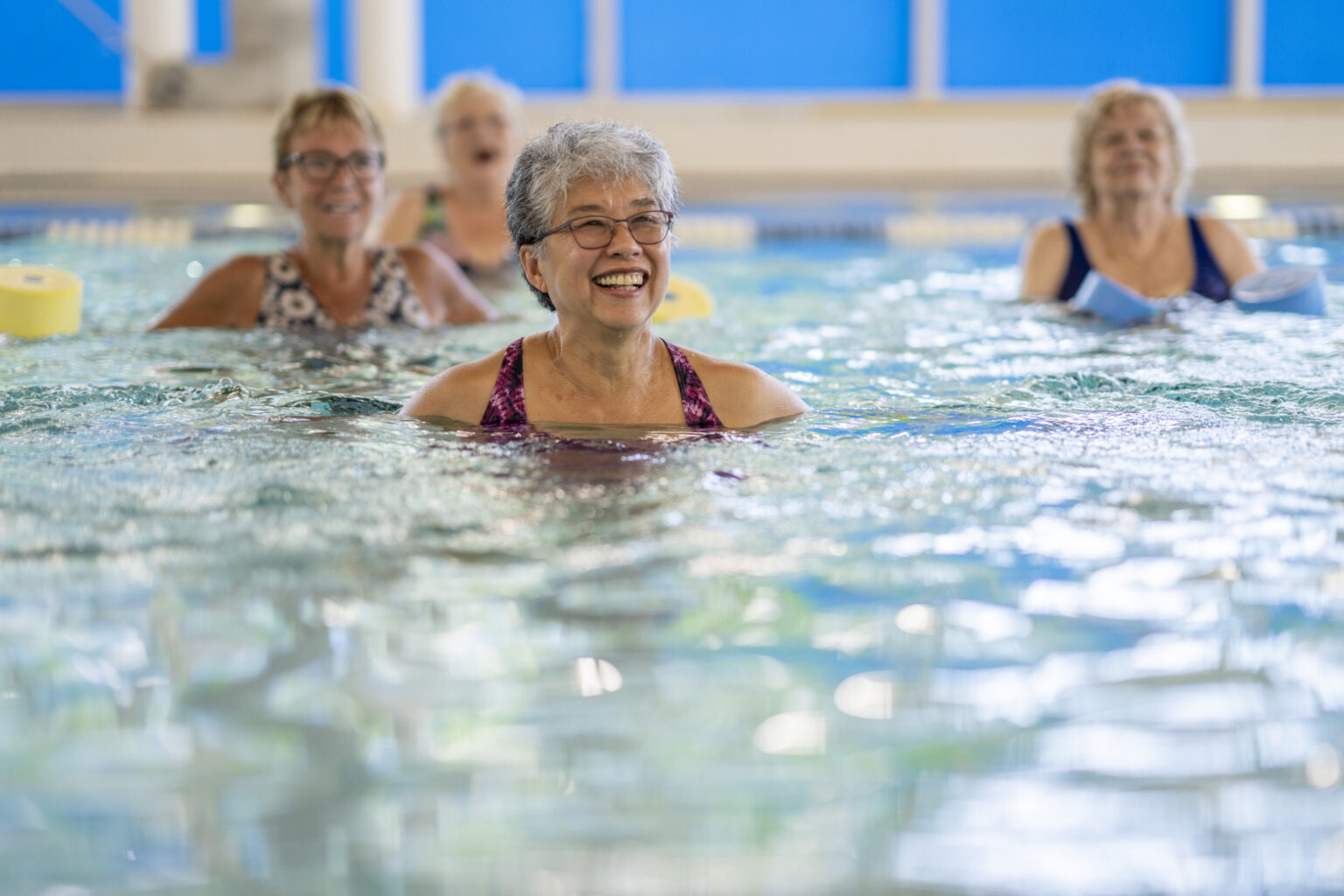 A senior woman is seen swimming in a pool as she laughs and enjoys the activity. She is wearing a swim suit and is among friends as she participates in an Aqua fitness class.